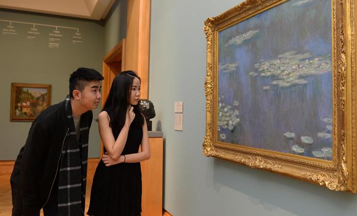 Young man and woman, both wearing black and with black hair. They are looking at a painting called Waterlilies, painted by Monet. Painting shows the b